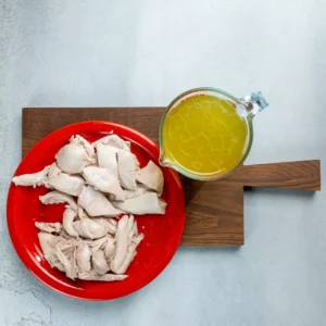 Ausgelöstes Hühnerfleisch auf einem Teller, daneben ein Krug mit Brühe